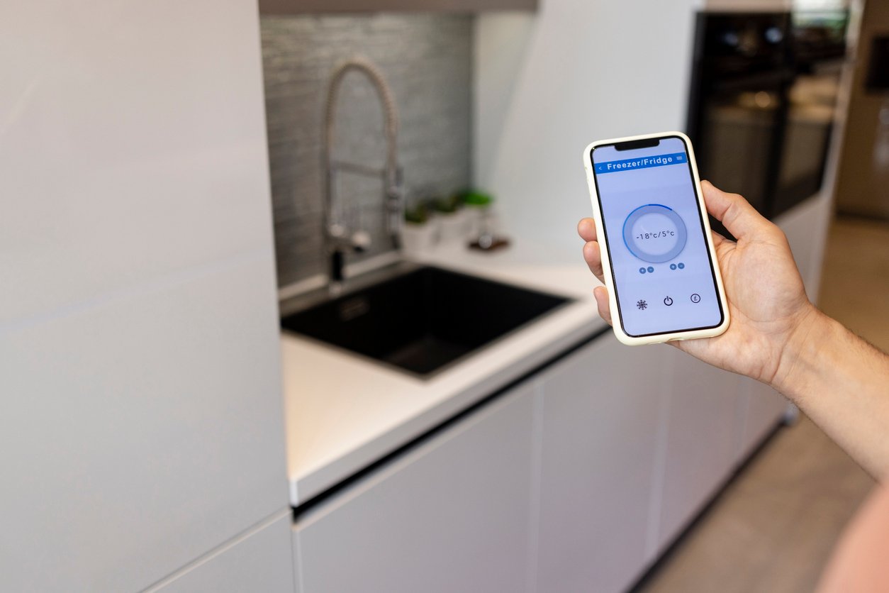 Hand holding a smartphone in a kitchen with a blurry image of a sink in the background, programming their energy-efficient faucets 
